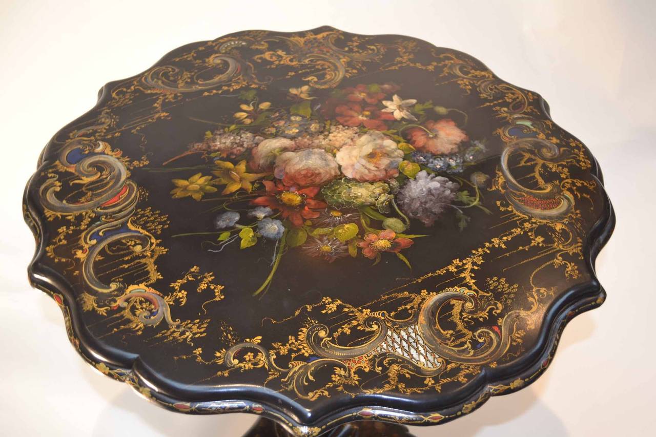 An English, (possibly French) papier mâché tilt-top table made, circa 1880. The entire table is ebonized and is richly decorated with gold leaf and painted flowers; the scalloped circular top is well painted with colorful bouquet of flowers and it