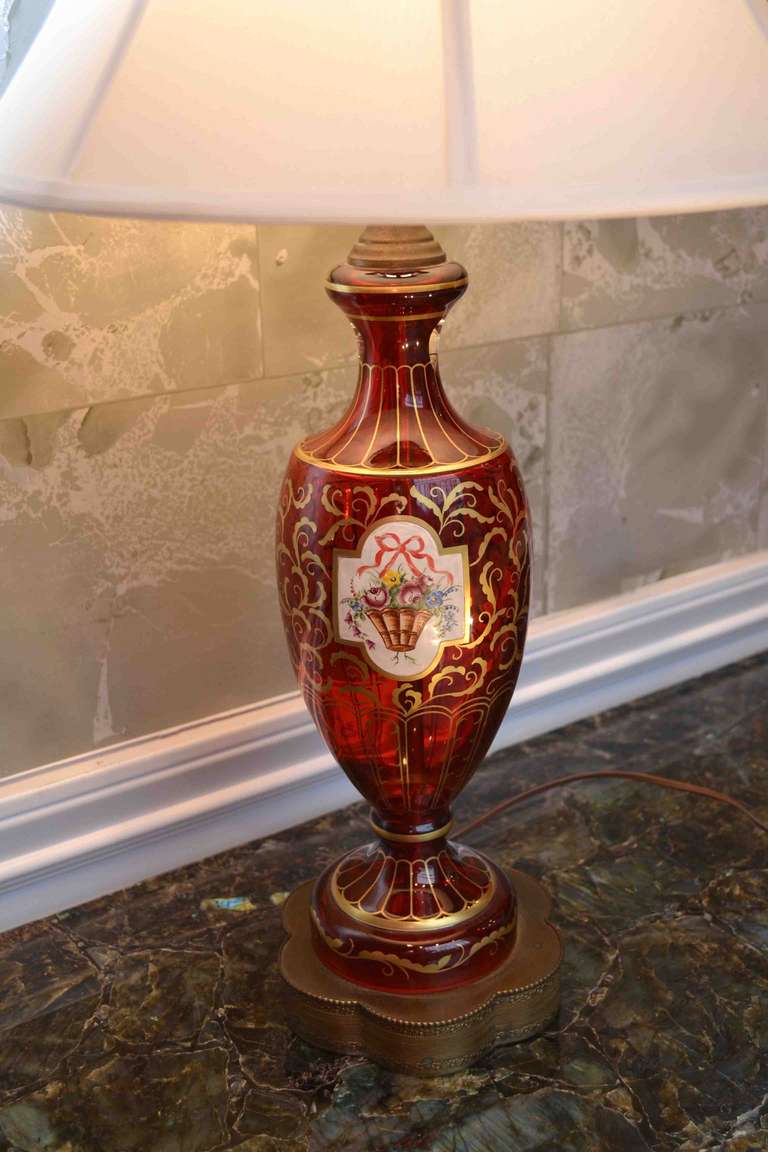 A classic urn shaped cranberry glass lamp decorated with gilding and a cartouche showing a painted Louis XVI style floral bouquet; mounted on a later gilded metal base.

Dimensions: 30