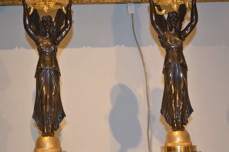 A very nice pair of gilt and patinated bronze candelabra of classic design; a patinated bronze figure of Nike stands on a gilded ball on an engine turned circular base and holds aloft a ring of gilded rosettes and oak leaves from which three candle
