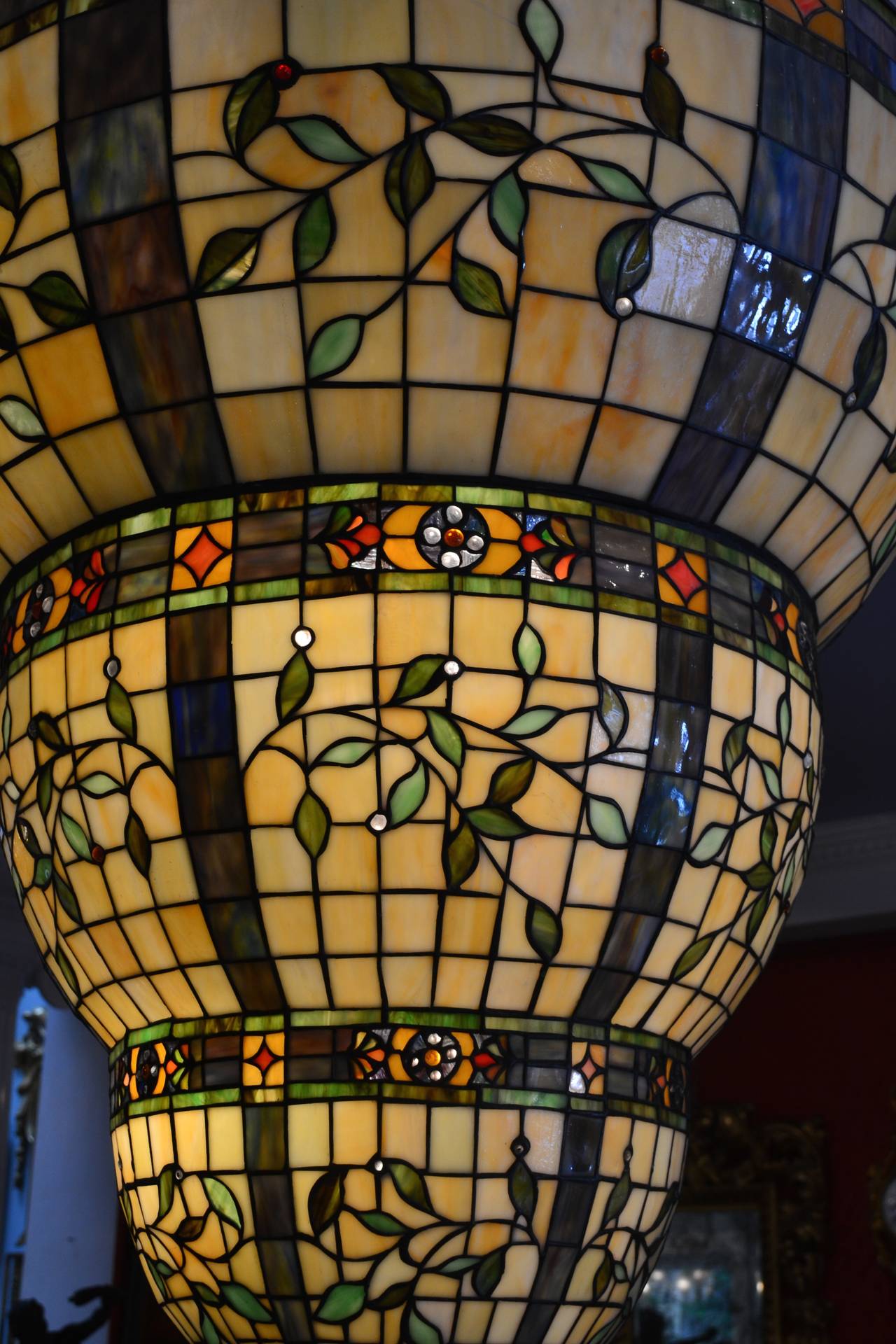 A wonderful ceiling fixture for the right locale. Three circular tiers of stained glass are suspended from six curved metal supports, each tier separated by a band of colorful rosettes and flowers, the tiers decorated in the round with branches and