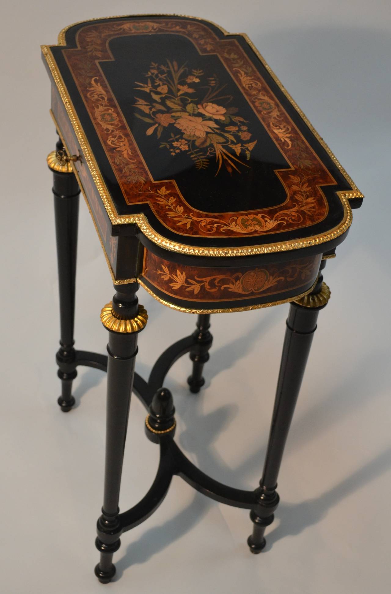 A very pretty sewing/occasional table having overall floral marquetry to the top and sides.  The rectangular shaped top with curved ends sits on four turned ebonized legs joined with a curved stretcher.  The table is further decorated with gilded
