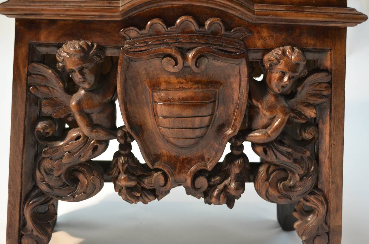 A nicely carved wooden stool/table, the wood stained to resemble walnut.  The front and back of the rectangular body are heavily carved showing two winged figures holding a heraldic shield, the two ends are also lavishly carved.