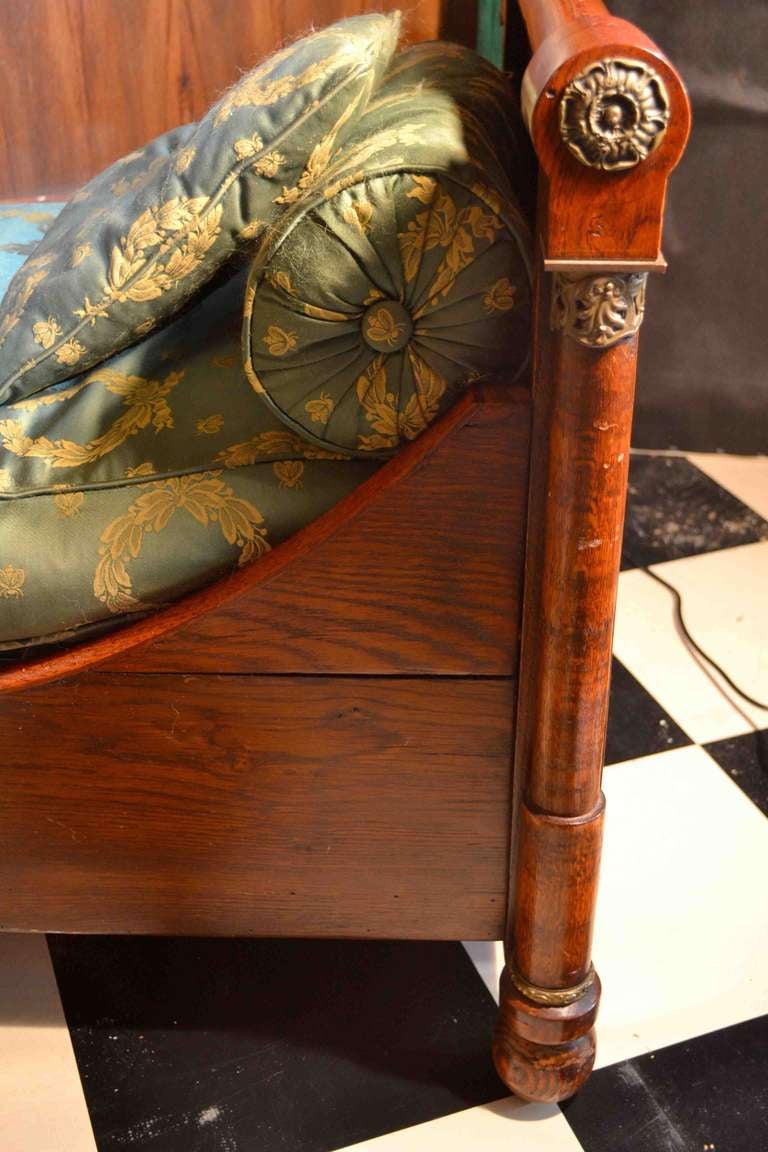 A small daybed/sofa in the full French Empire lit-de-bateau style, the oak frame stained and polished in a mahogany colour;  the end columns with gilded bronze feet and capitals; the seat, bolsters, and cushions upholstered in an Empire silk fabric;