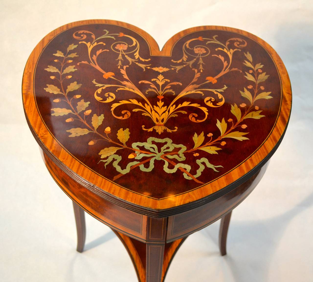 A unusual heart shaped table in mahogany; the top banded in satinwood and inlaid with various exotic woods in the shape of garlands, laurel and berries, and arabesques. Below the top are two 'secret' drawers that open sideways revealing velvet lined