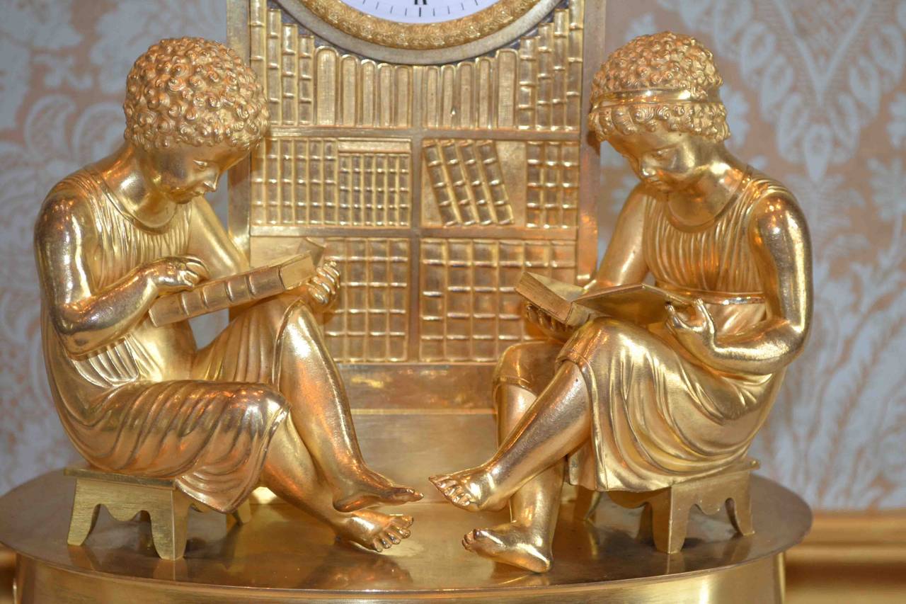 A fine and rare French Empire clock in gilded bronze. Two boys dressed in classical attire are diligently studying in a library, both are sitting in front of a clock which is integrated into a column which also serves as a bookshelf. The clock base