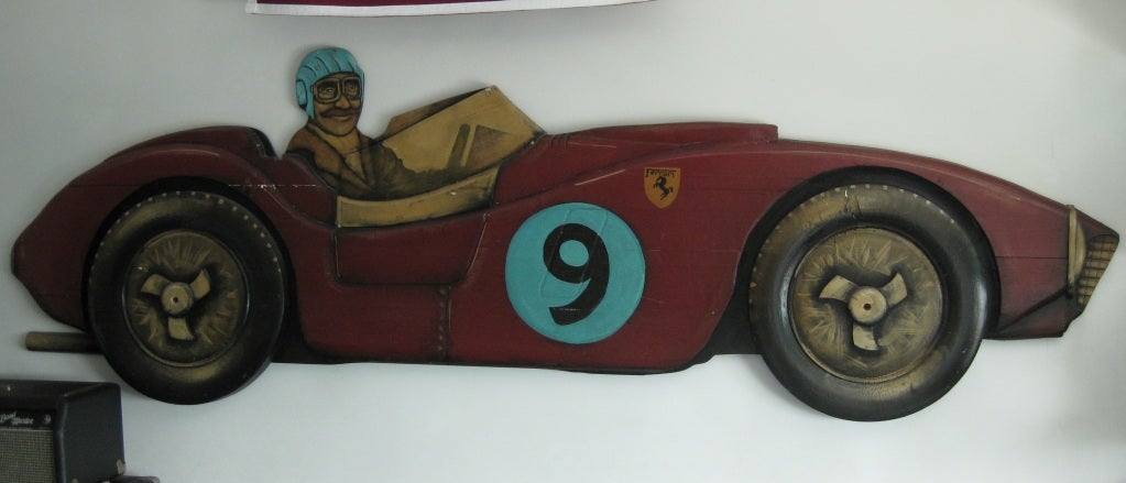 This unusual carved and hand-painted wood bas-relief depicts a driver with aviator jacket, helmet, goggles and ascot seated in his winning car; one of the most famous cars ever designed by Enzo Ferrari, a 1957 Ferrari Testarossa. The sculpture can