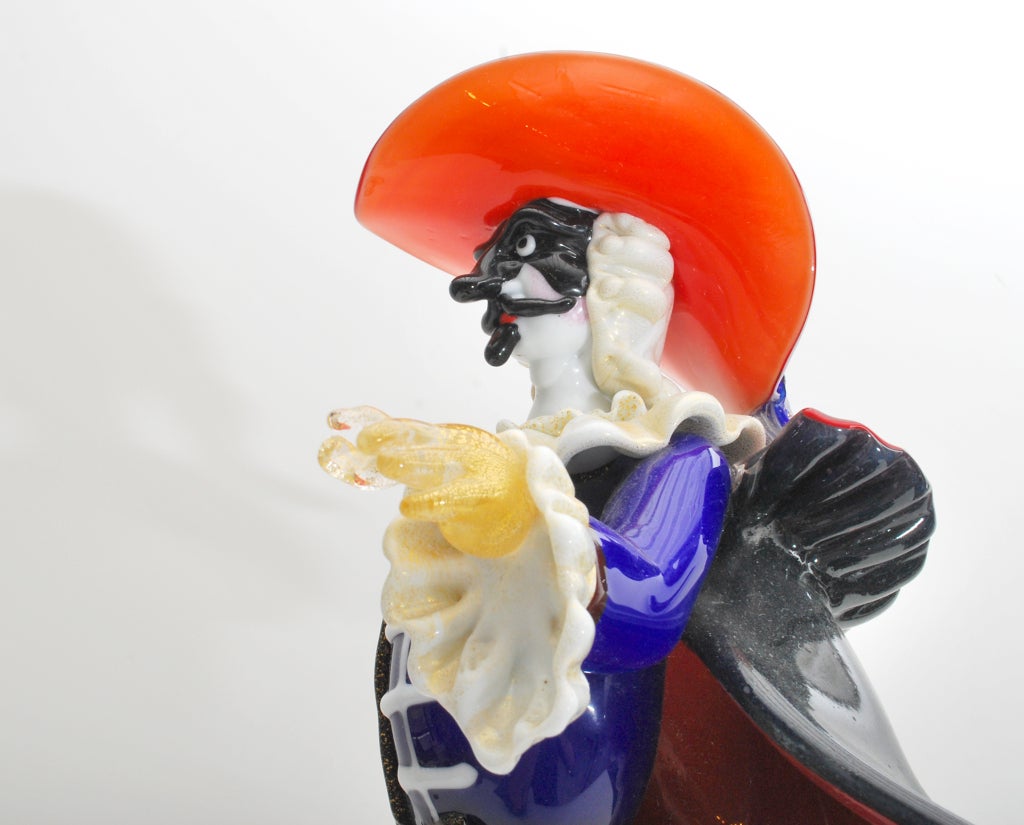 Signed Pino Signoretto figurine of a masked musketeer in colorful 17th century costume with flowing cape standing on a base with lion head prunts, Murano, Italy. *Notes: There is no sales tax on this item if it is being shipped out of the state of