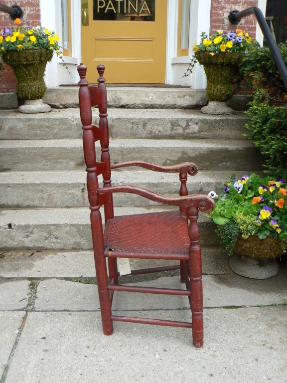 Remarkably preserved 4-stretcher ladder-back chair in original red paint. Turned and incised legs and back with knob finials. Bulbous front stretcher. Arched arms terminate in uniquely shaped hand rests. Woven Seat.