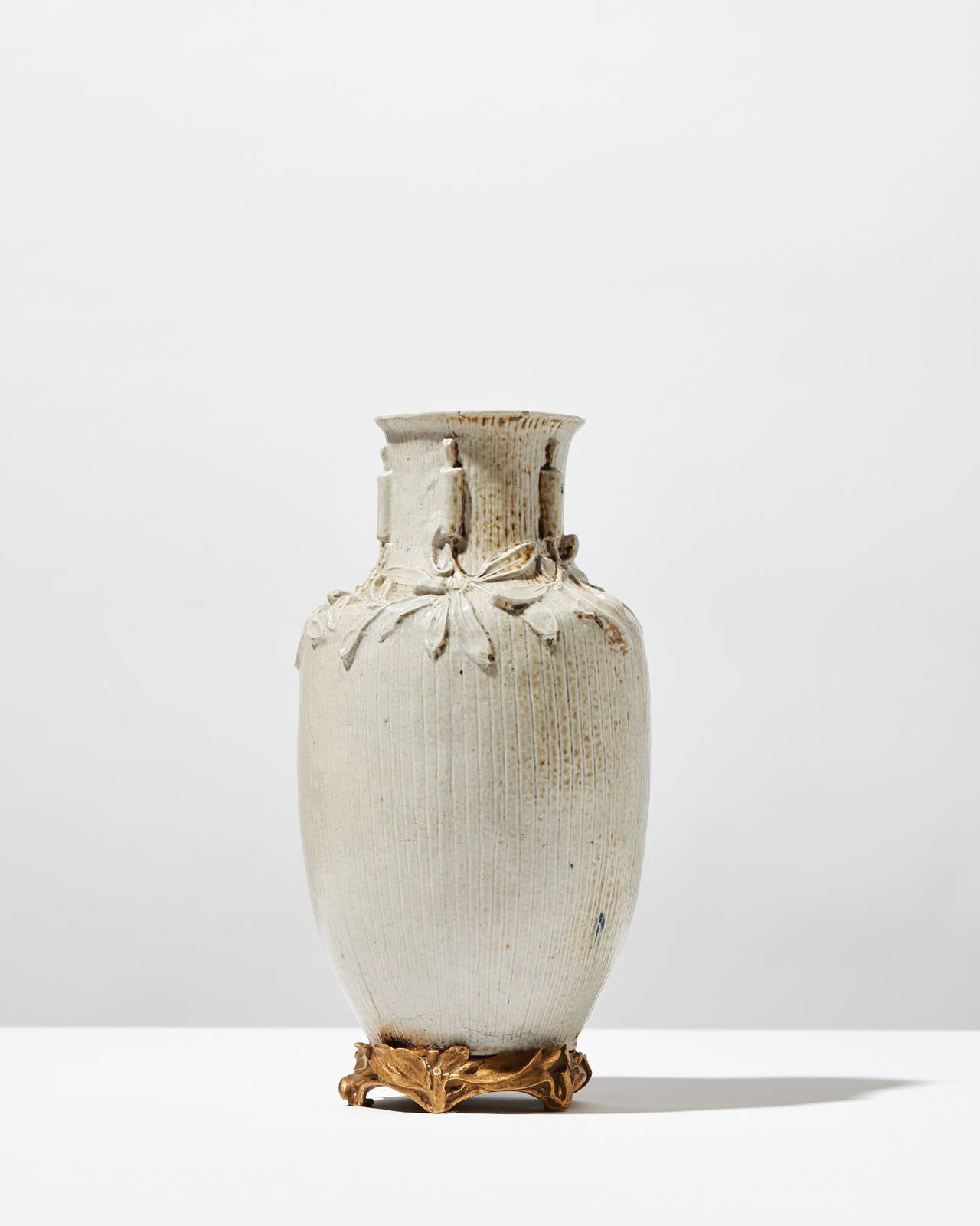 This vase rests upon a bronze mount, which contrasts with the vase's rather matte white finish. The vertical indentations point upwards towards the narrowed neck and flared mouth. Gathered near the neck are a series of plants that are hung upside