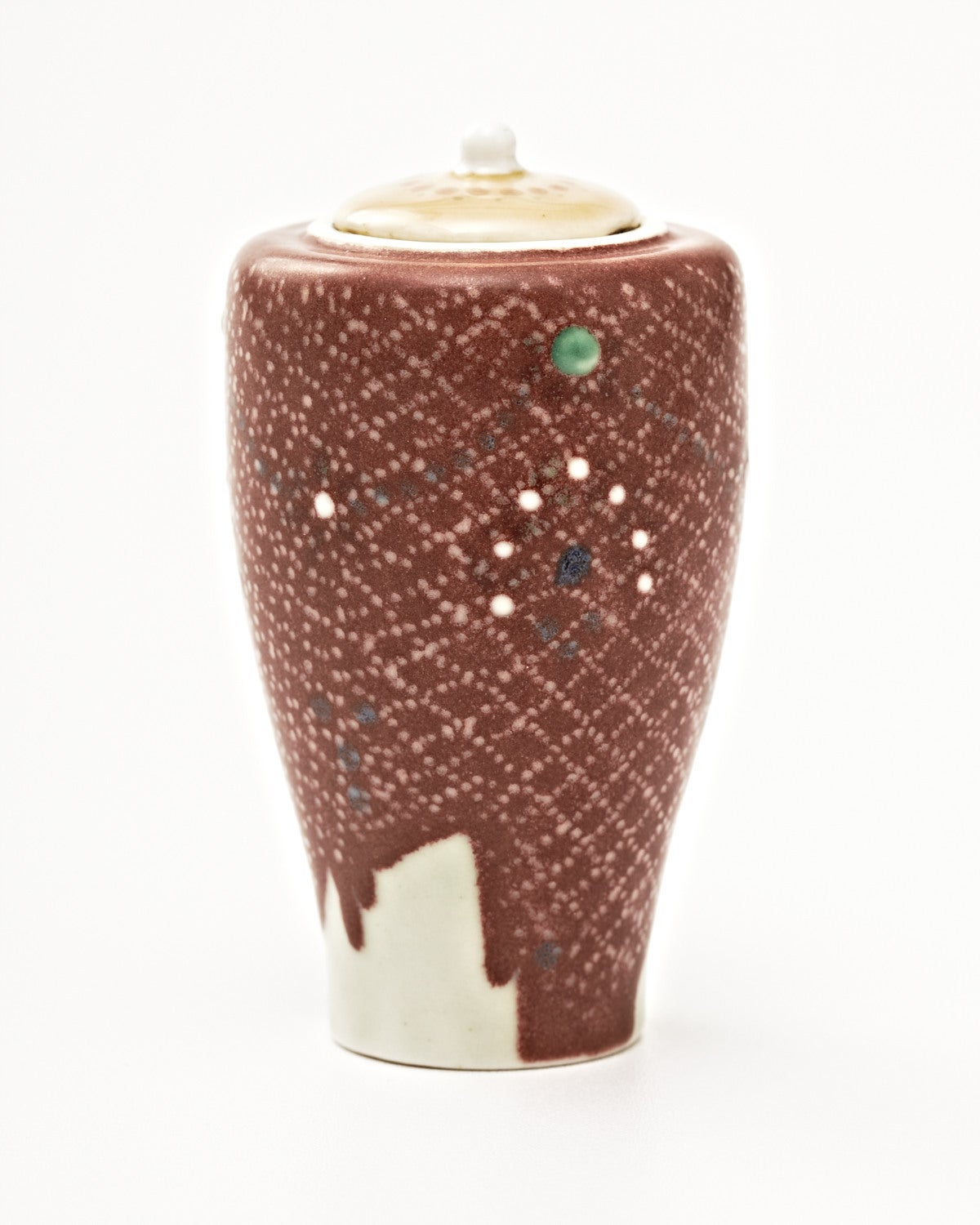 This petite lidded vase of elegant tapering form, is richly decorated with a thick dripping brown glaze which is covered in a delicately rendered, pointillistic grid-like a pattern. Further adorned with spots of green, white, and dark blue, the