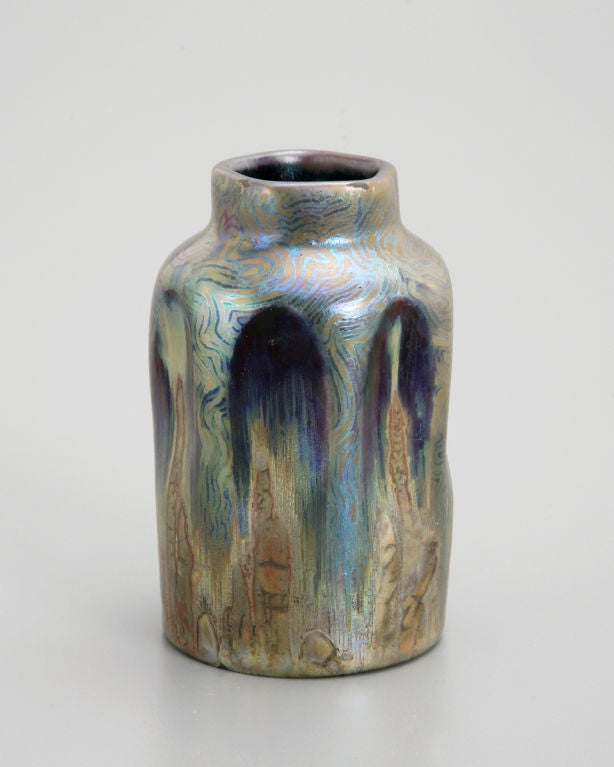 The undulating surface of this diminutive vase creates arch-like motifs that are ornamented with wavy lines of iridescent purple and gold. Dark purple pools beneath the arches suggest depth and distance, while in the foreground, ochre seaweed