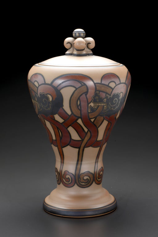 This broad-shouldered container with flaring foot and circular lid, features stylized interlaces with biting dragon heads in black and brown on an ochre ground. Interlacing dragon designs of this type originated in the medieval wooden churches of