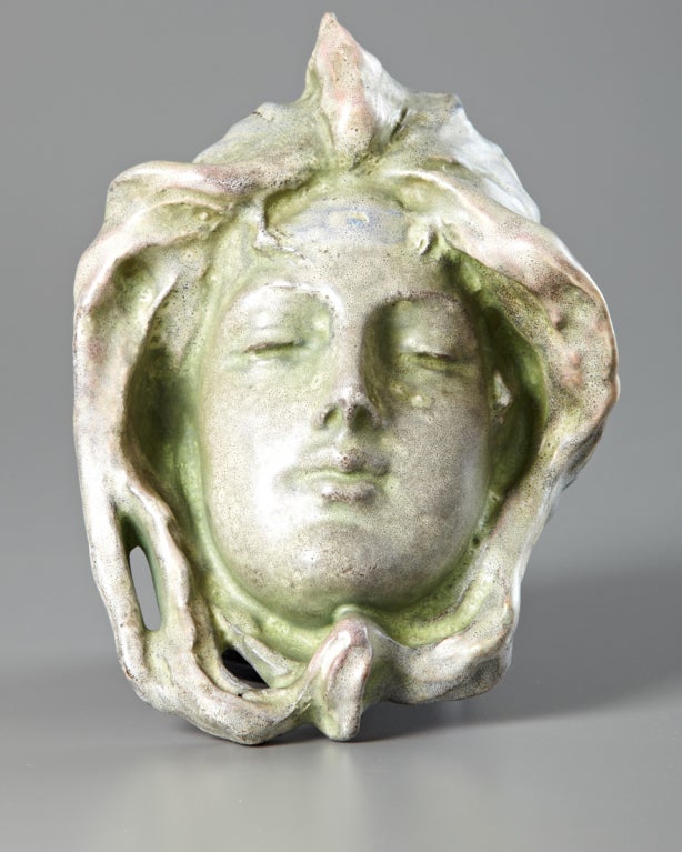 The serene expression of the maiden depicted in this stunning earthenware wall mask exudes a certain ghostly beauty. Decorated in a remarkable mottled matte glaze in a greenish grey hue, with pale pink and blue accents, this fine ceramic sculpture