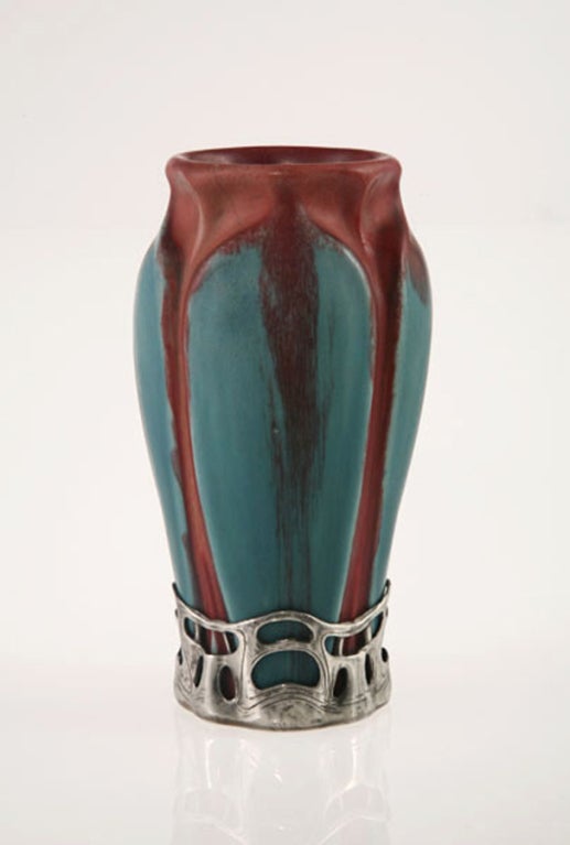 This sculptural vase is clad in a glaze of blue-turquoise with deep maroon around neck and in steaks from neck to base. Its preciousness is enhanced by a fine metal mount at the base.