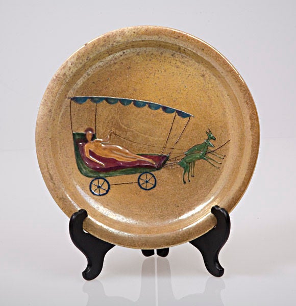 An earthenware charger with a whimsical decor depicting a female nude reclining in a cart drawn by antelopes. The decor is executed in enamel and luster glazes in low relief.