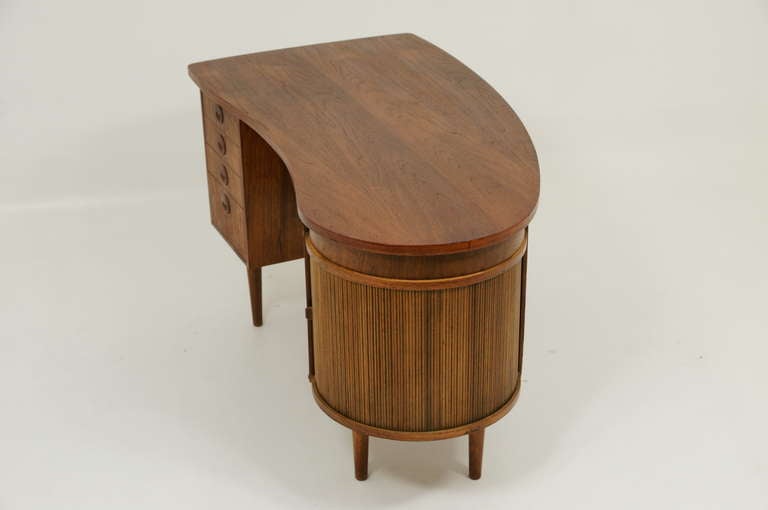 Gorgeous teak desk from Denmark with a very unusual shape. Drawers down one side and a very cool tambour door section on the other side that opens to show a revolving bar with mirrored back. Back side of desk has an open shelf and a large