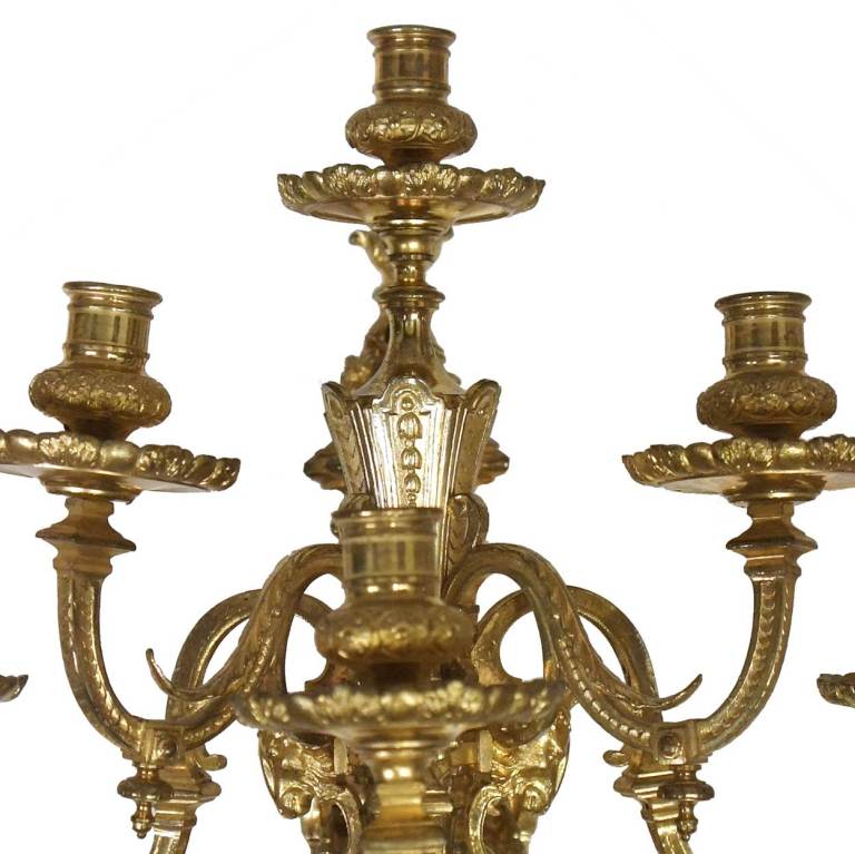 In the style of the 18th century, these wall lights are cast with a  backplate supporting a quiver form stem with rams head mounts. The stem is topped by a drip pan and candle nozzle, and issues four S-scroll arms.
