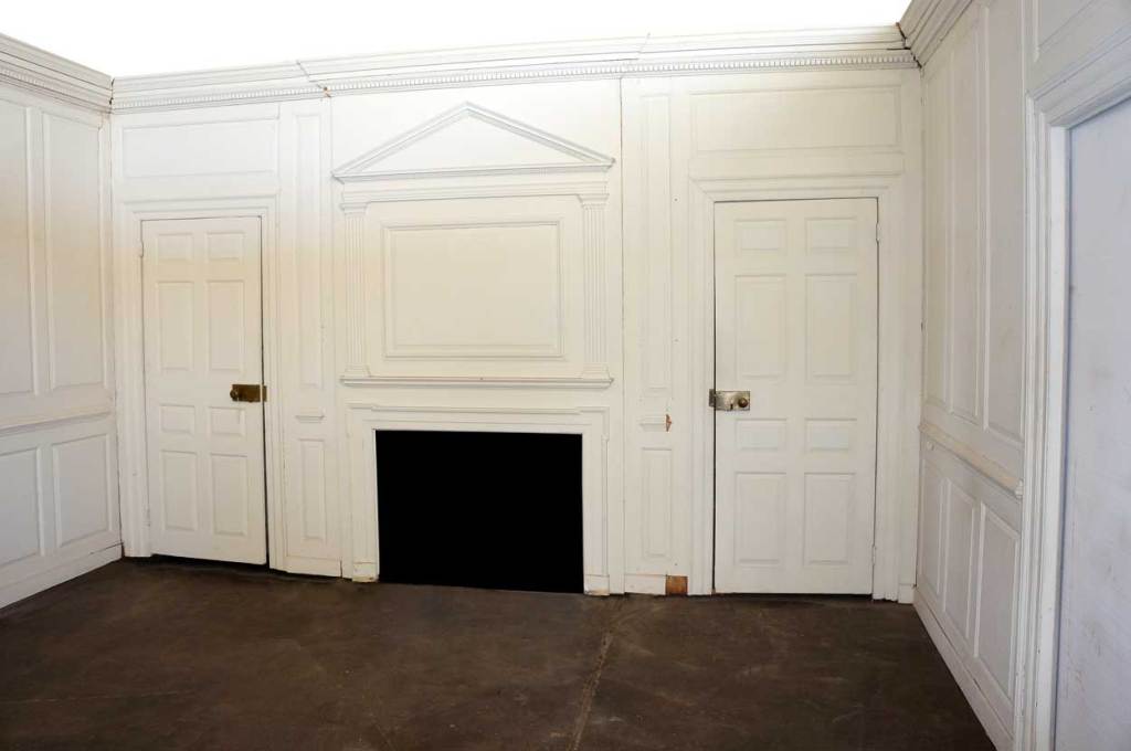 This elegant period room includes dentil crown moulding, a fireplace surround with a neoclassical overmantel with fluted pilasters and pediment, doors, fielded panel walls, mullion windows and window seats. The white paint is a later addition.