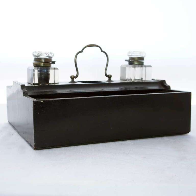 This ink stand has a rectangular two-tier frame with reeded edges and a brass carrying handle. It has three square wells for ink bottles, over two pen trays, one with a sliding lid and brass knob, above a long base drawer on one side. Together with