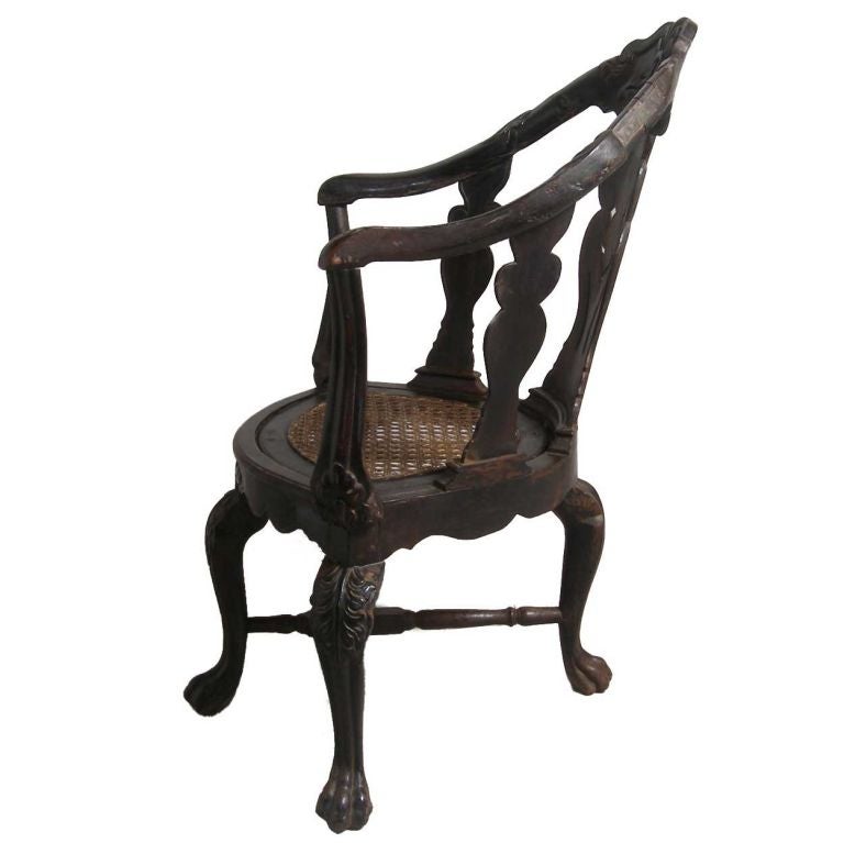 This elegant chair was handcrafted in Goa. It displays a concave, carved crestrail with downward scrolling arms with a pierced splat. The caned seat has a serpentine apron and rests on four cabriole legs with paw feet joined by a turned x-stretcher.