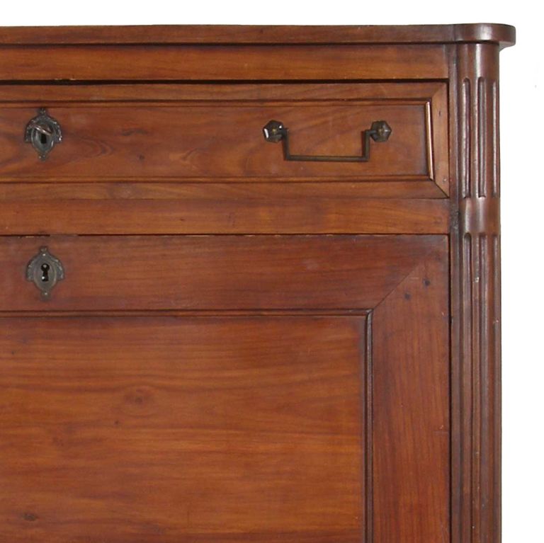 This desk cabinet has a rectangular top with eared corners with a conforming case fitted with a frieze drawer with brass, bail handles, above a drop front desk, lower cabinet doors and tall tapered legs. The interior has an arrangement of arched