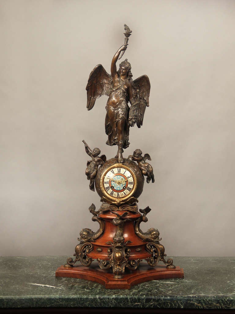 Grande et importante horloge de cheminée en bronze doré et patiné, en émail cloisonné champlevé et en bois de la fin du XIXe siècle.

La figure du haut représente Nike de Samothrace, la déesse grecque de la victoire. Deux chérubins représentant