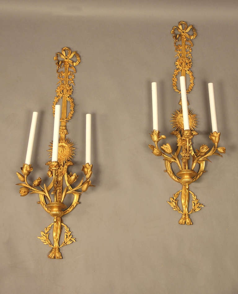 A superb pair of late 19th century Louis XVI style gilt bronze three-light sconces.

The backplate formed as a bow knotted ribbon continuing to a floral wreath, a sun-god mask and a lyre issuing floral candle branches.