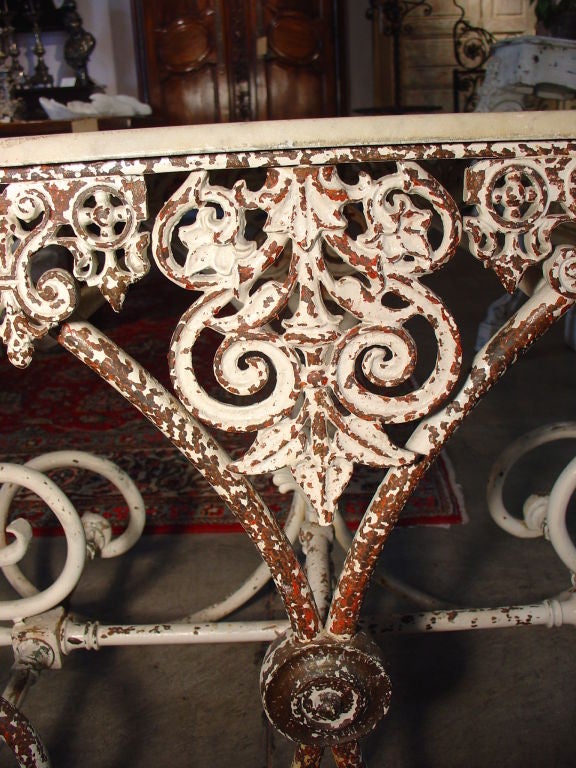 his lovely antique French pastry table has a marble top with a painted iron base.  The apron has stylized fleur-de-lys formed with c-and s-scrolls and a central volute with fleur-de-lys drop.  The legs are large c-scrolls on either side connected