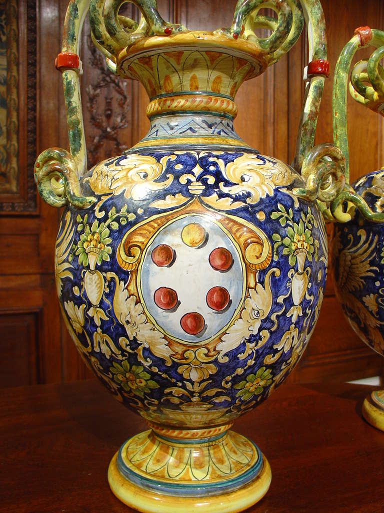 This pair of colorful hand painted Italian urns has stylized serpent handles with bodies that have different cartouches on either side with different motifs.  The ground colors on both are a beautiful deep cobalt blue with areas of white, cream,
