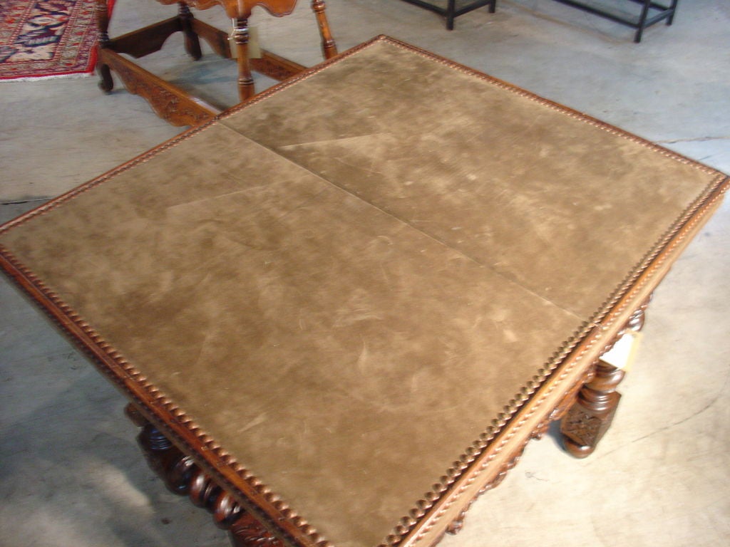 This antique French Louis XIII style games table has a fold out top that is covered with a faux suede material affixed to the wooden top with brass nail heads.  There are square wooden pieces which slide out to hold the extended top on either side. 