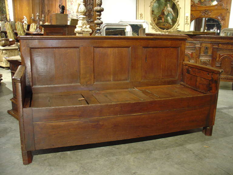 This antique  wooden bench from France has a three paneled back with paneled side arms and a seat with storage beneath it.  There are hinged leather tabbed panels which open to the storage below. The front of the seating area is a plain panel. 