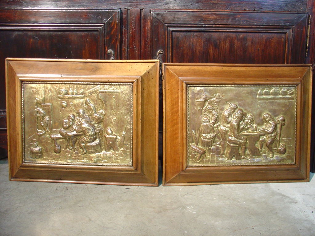 This charming pair of antique French brass repousse framed plaques features festive scenes of people in rooms having ancient stone floors, fireplaces, and various pieces of antique furniture and accessories typical of a very old French