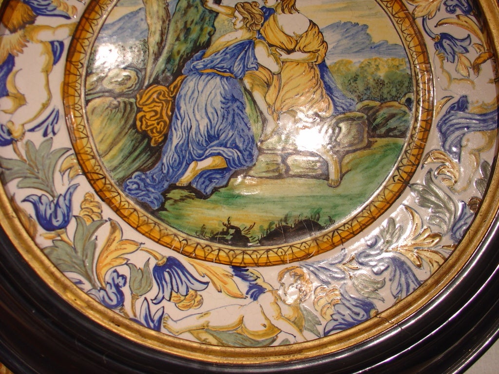 This high fired, glazed faience has a high gloss thick black frame surrounding it. The scene is that of a man and a maiden overlooking the countryside. The colors are numerous on this piece. Surrounding the scene, is a border of floral foliage,