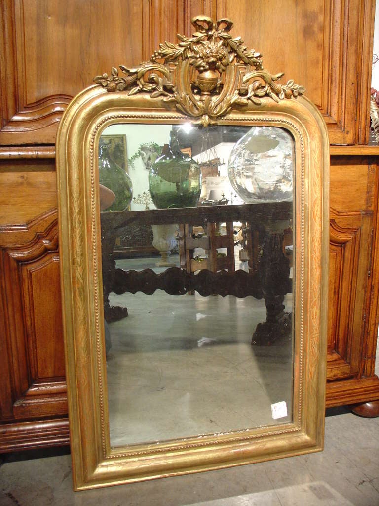This antique French Louis Philippe mirror has a cartouche motif at the top center.  There are motifs of a tied ribbon holding sprays of laurel leaves and berries, and a vase filled with floral and foliate motifs.  This is surrounded by fretwork with