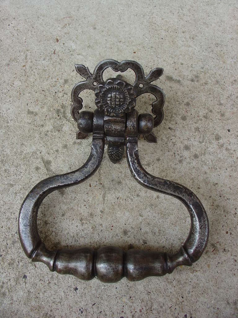 This rare antique French hand forged iron door knocker is from the 1700’s.  At the top of the knocker is a primitive floral motif with a patterned ground at its center.  At the other end of the flower is a motif of a nutmeg.  The back plate has open