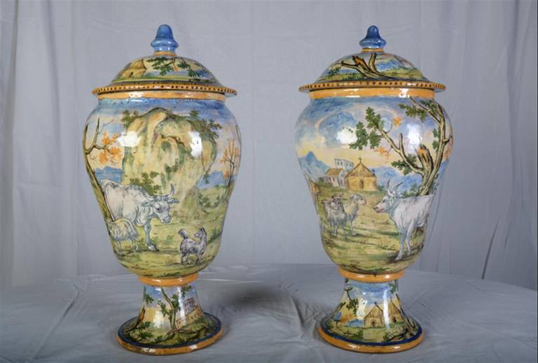 These magnificent Italian Majolica lidded vases are hand painted . The Genre painting depicts a continuous scene of what would have been  an urban setting in the 1700’s. The people are depicted with their animals including goats, pigs, chickens,