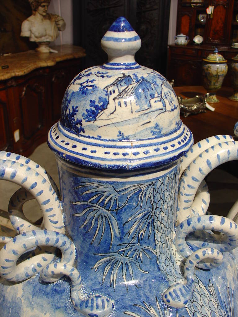 This magnificent and large blue and white Savona type lidded Majolica urn has hand-painted scenes on both sides. The handles depict serpents 'bodies circling outward and ending over two bas relief mascarons' faces. Toward the bottom of the 'front'