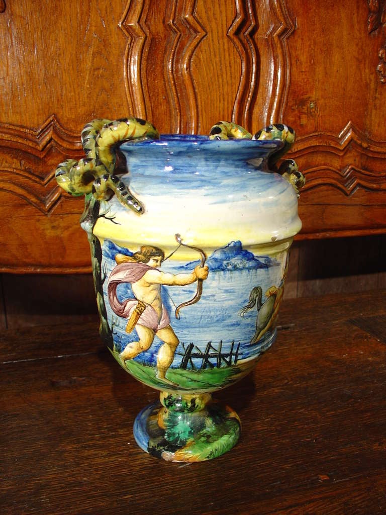 This antique hand painted tin-glazed Majolica urn was done at the famous ceramics factory of Ulisse Cantagalli, near Florence.  The baluster shaped urn has curled serpent handles and exhibits a continuous scene of an outdoor Roman setting.  There is