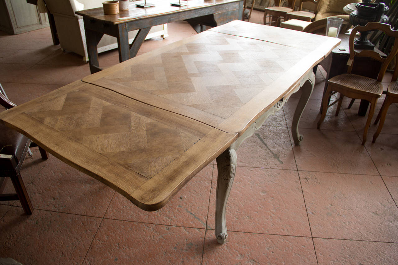 This carved antique Louis XV revival draw-leaf oak table from France has an inlaid parquetry top on a painted base. The top sits on cabriole legs with escargot feet and beautiful scroll and anthemion carvings.

It extends to 7.4 ft.