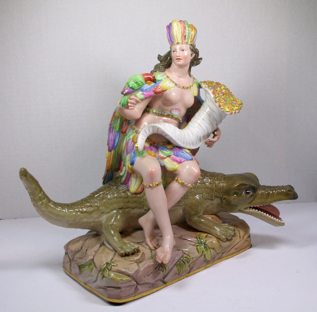 Allegorical porcelain figure of America from the Continents series in the form of a woman wearing a brightly feathered outfit, holding a parrot and gem filled cornucopia<br />
and seated on an alligator.  The Continents series was created by the
