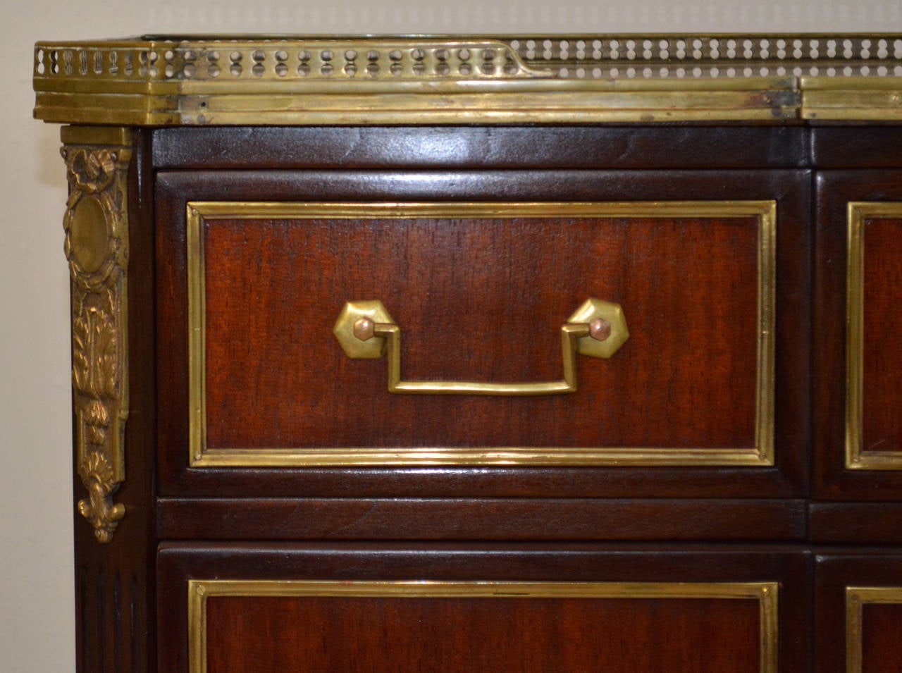 A lovely three drawer commode made of light and dark stained mahogany with brass mounts in the style of Maison Jansen's early 20th century work. Drawers feature brass trim, key escutcheons and classic drop handles with hexagonal backplates. A white