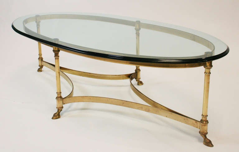 Lovely oval cocktail table with glass top. Brass base with four legs, hoof feet, and curved stretchers. In the style of Maison Jansen.