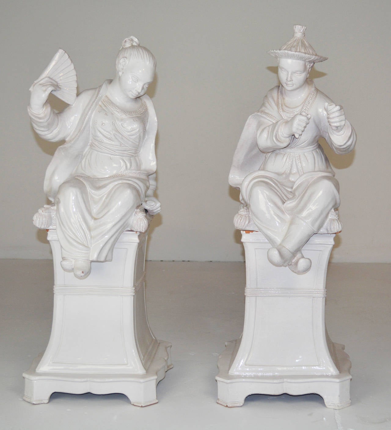 An interesting pair of large scale Italian white glazed terracotta figures of a Chinese girl with a fan and a boy, each seated on a pillow and mounted on its original matching pedestal with flared base and incised panel. Marked on underside Italy.