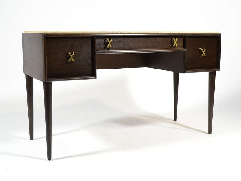 This design by Paul Frankl is has great scale and versatility. It is useful as a writing desk, vanity, or console table. It features his signature X-shaped brass drawer pulls, two deep drawers, one shallow drawer, and sits atop four tapered legs.