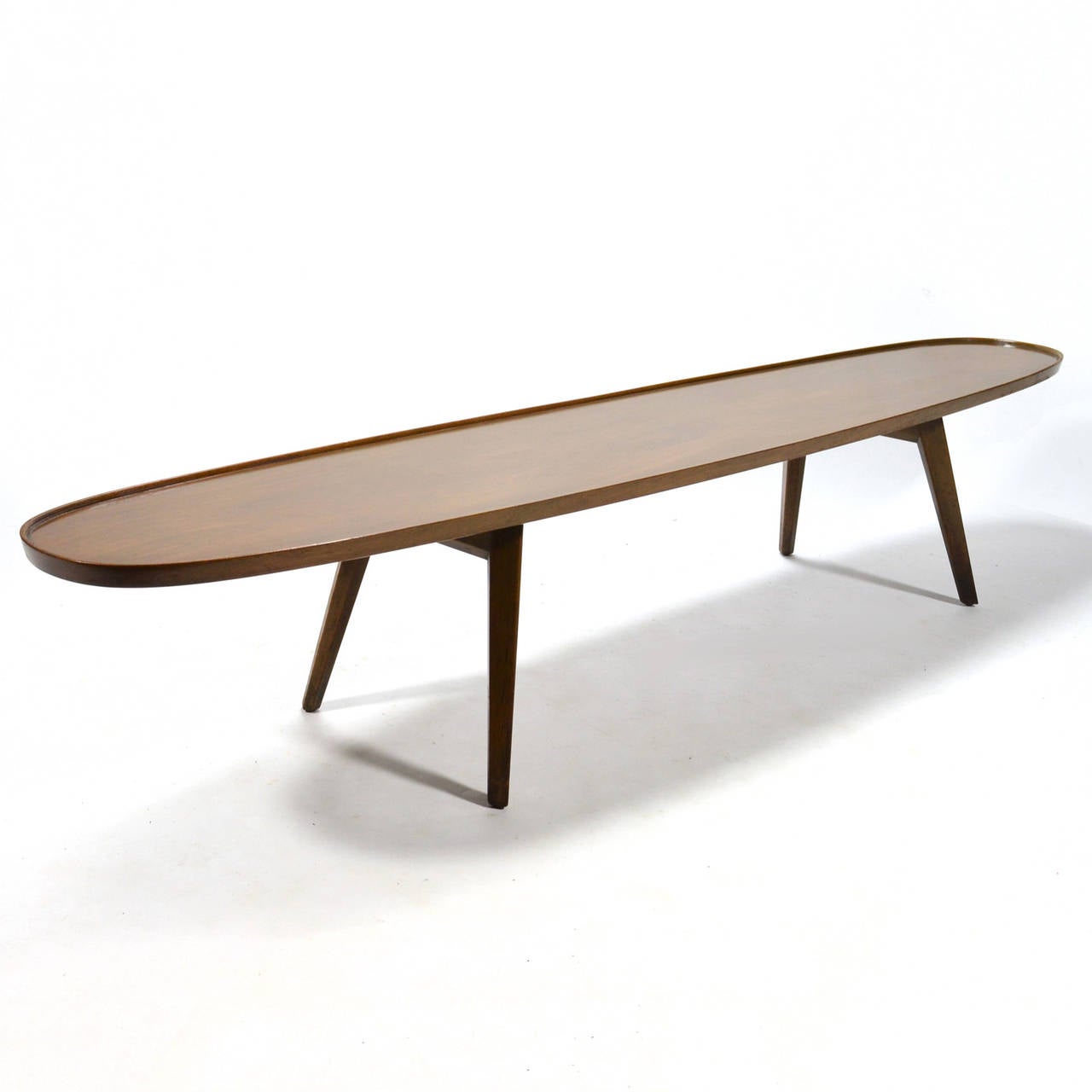 An early Edward Wormley design for Dunbar, this beautiful long walnut coffee table has the elongated elliptical shape of a vintage surfboard. It is supported by four splayed tapering legs with faceted sides and the top has a thin raised pie crust