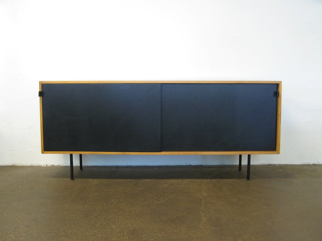 This handsome credenza designed by Florence Knoll (model 116) has a maple case with two sliding doors of black leather. The doors have leather tab pulls and conceal 4 bays with adjustable shelves and a divided drawer. The cabinet is supported by