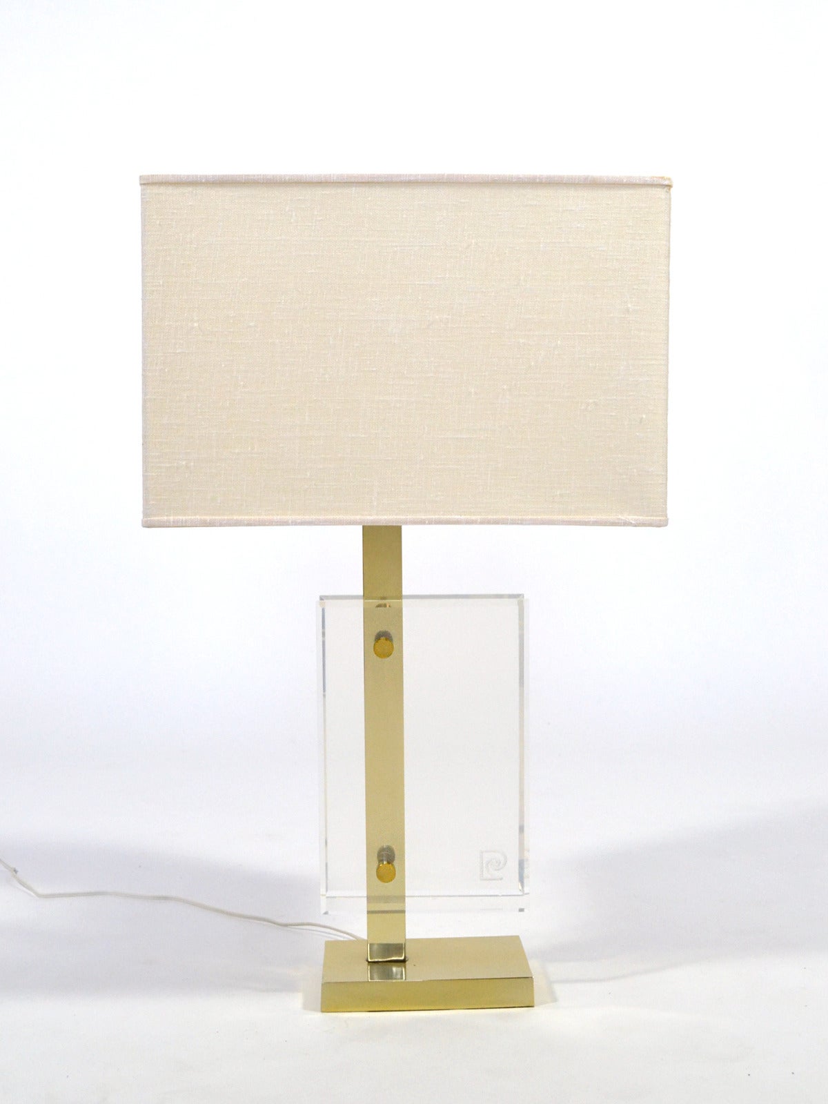 A terrific asymmetrical design, this Pierre Cardin table lamp has a base of brass with a pair of Lucite forms, one etched with the Cardin logo. It retains the original rectangular linen shade. Like Cardin's fashion designs, the lamp is elegant, sexy