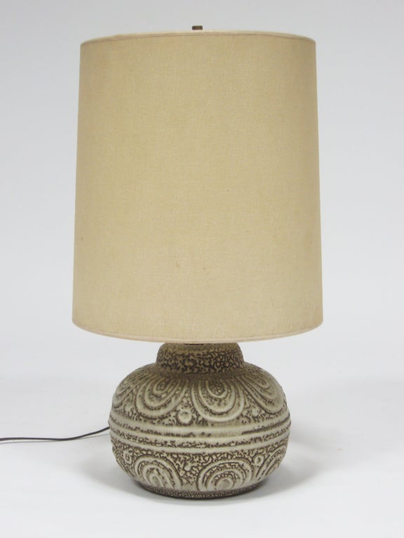 A delightful design, this table lamp features a ceramic base that has a richly textured and patterned surface. It is topped with a tall cylindrical shade that creates a wonderful tension between the tall, smooth fabric shade and the short, nubby