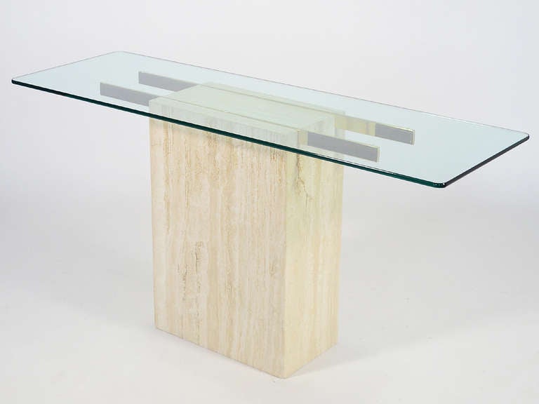 An elegant minimalist form, this console table by Ello has a base of Italian travertine marble and a pair of brass arms which supports  a glass top. The monolithic stone base is the perfect counterpoint to the transparent glass and light linear arms.