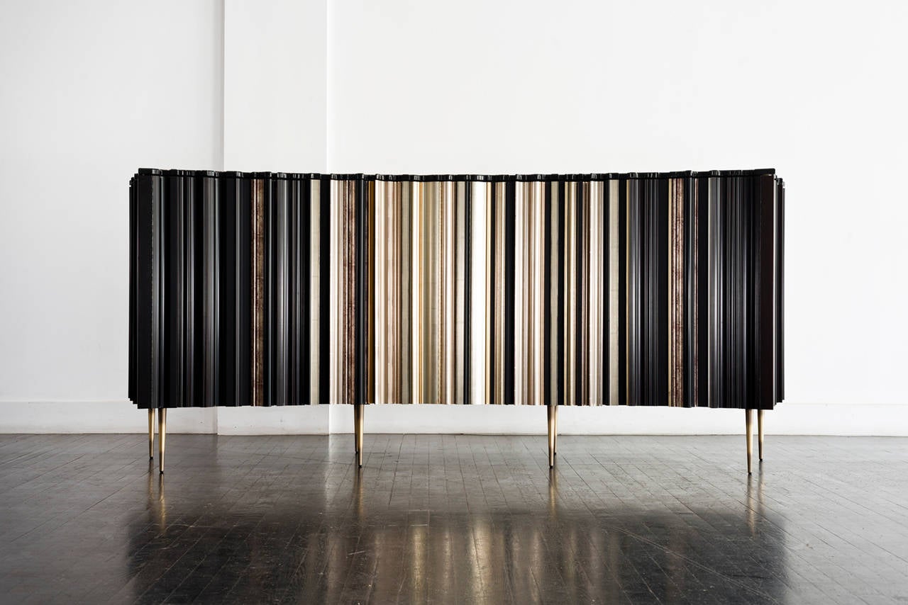 With its elegant concave face, this striking cabinet has individually selected frames applied vertically to a wooden case with tapered brass legs. The graphic pattern of the dark and gilded frames is unique to each piece. 
The black lacquered top